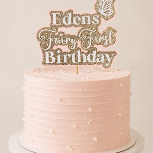 May include: A light pink cake decorated with small white pearls and a gold glitter cake topper that reads Edens Fairy First Birthday with a white butterfly icon. The topper is mounted on a thin wooden stick inserted into the cake top.