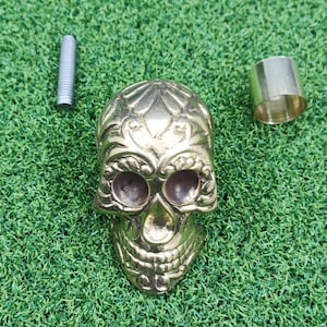 May include: A decorative brass skull with detailed floral designs, alongside a metal bolt and a cylindrical metal component. The skull is the central element, set against a green, textured backdrop.