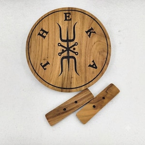 May include: A circular wooden board with a natural grain finish, featuring a black engraved trident-like symbol in the center and the letters H, E, K, A arranged around it. Two separate rectangular wooden pieces with pre-drilled holes are included.