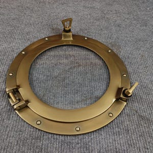 以下が含まれることがあります： 真鍮色の丸い舷窓で、ヒンジ付きの開閉機構と目に見えるリベットが付いています。舷窓には開閉用のハンドルが付いています。航海をテーマにしたアイテムは、灰色の表面に置かれています。直径約25cm。