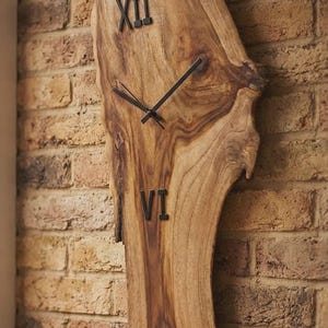 Puede incluir: Un reloj de pared fabricado con una pieza de madera natural con borde vivo. La esfera presenta una veta de madera pronunciada, manecillas de metal negro y dos números romanos negros que representan el doce y el seis arriba y abajo.