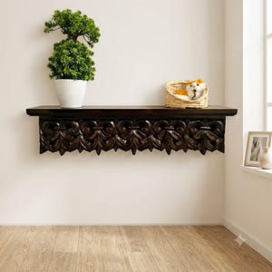 Peut inclure: Une étagère murale en bois brun foncé avec un motif floral sculpté complexe sur le bord avant. Une plante verte dans un pot blanc est posée sur le côté gauche, et une petite peluche blanche et orange dans un panier en osier est sur le côté droit.