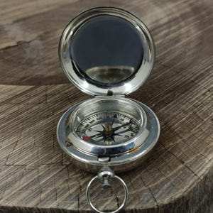 May include: A silver compass with a hinged lid and a ring for attachment. The compass face features directional markings and a needle. The open lid reveals a reflective surface. The compass is set on a wooden surface.