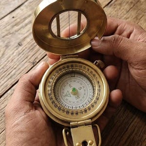 May include: A vintage brass compass with a hinged lid, held open to reveal the compass face. The compass has a detailed dial with directional markings and a needle pointing north. The brass casing has a polished finish.