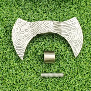 以下が含まれることがあります： 木目模様の銀色の両刃の斧頭、銀色の金属シリンダー、銀色のねじ付きボルトが緑色の表面に置かれています。 斧頭は湾曲した形状をしています。