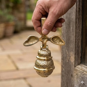 Può includere: Un battente per porta in ottone dal design unico. Presenta una maniglia decorativa con elementi a forma di ala e un corpo arrotondato e strutturato. Il battente è tenuto in mano, suggerendo il suo utilizzo per una porta.