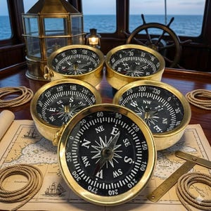 May include: Five antique brass compasses with black faces and white markings are arranged on a wooden surface with a nautical map. The compasses are surrounded by nautical ropes and a brass divider. A ship's wheel is visible in the background.