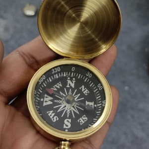 May include: A vintage brass compass with a hinged lid, open to reveal the directional dial. The compass face is black with white markings and a needle pointing north. A small ring is attached for carrying.