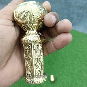 May include: A gold-colored, ornate cane handle with intricate floral and leaf patterns. The handle has a rounded top and a rectangular base, held in a hand against a green and blue background. The handle is made of metal.