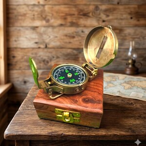 May include: A vintage brass compass with a green and black dial, open on a wooden box. The compass has a hinged lid and is set against a wooden background. The box is rectangular with a brass clasp.