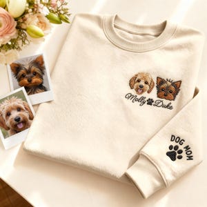 Op de afbeelding: Crèmekleurige sweatshirt met geborduurde hondenportretten en de namen "Molly" en "Duke". De mouw toont "DOG MOM" en een pootafdruk. Twee foto's van honden zijn naast de sweatshirt te zien.