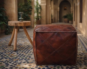 Può includere: Un pouf in pelle marrone con un motivo a rombi, posto su un pavimento piastrellato. Un tavolino in legno con piano rotondo e tre gambe è accanto al pouf. La scena è ambientata in un cortile esterno.