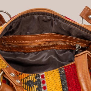 Op de afbeelding: Detailopname van een kleurrijke handtas met een patroon in rood, geel en zwart. De tas heeft bruine leren accenten, waaronder handvatten en een ritssluiting. De binnenkant is gevoerd met een donkere stof.