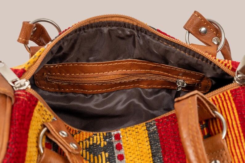 Op de afbeelding: Detailopname van een kleurrijke handtas met een patroon in rood, geel en zwart. De tas heeft bruine leren accenten, waaronder handvatten en een ritssluiting. De binnenkant is gevoerd met een donkere stof.