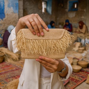 May include: A rectangular, woven straw clutch with a fringed edge. The clutch is held up by a person wearing a white blazer. The background shows a workshop with other woven items and people working on them.