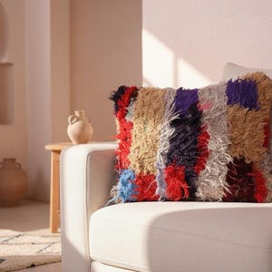 May include: A colorful, textured pillow with a patchwork design rests on a white sofa. The pillow features a mix of red, blue, purple, and tan hues, creating a vibrant accent piece for home decor. The pillow's shaggy texture adds visual interest.