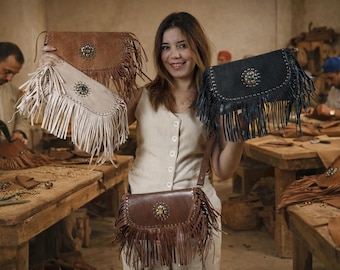 Può includere: Una donna mostra diverse borse con frange in pelle di vari colori: marrone, crema e nero. Ogni borsa presenta un elemento centrale decorativo e una lunga tracolla. Le borse sono fatte a mano, con artigiani che lavorano sullo sfondo.