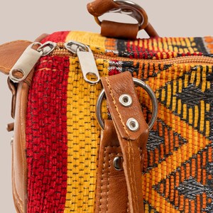 Op de afbeelding: Close-up van een kleurrijke tas met een geweven patroon in rood, geel en zwart. De tas heeft bruine lederen accenten, zilveren ritsen en een stevige handgreep. Het ontwerp bevat geometrische vormen.