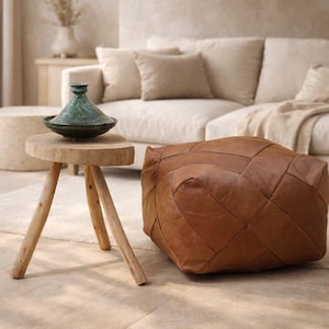 May include: A brown leather ottoman with a geometric design is on a light floor. A small, round wooden side table with three legs holds a green ceramic tagine. A beige sofa and rug are in the background.