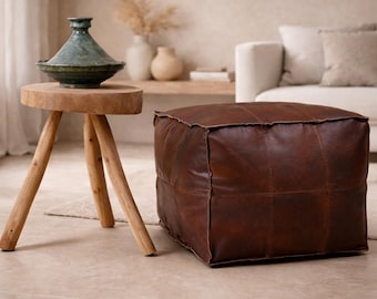 Può includere: Un pouf in pelle marrone di forma quadrata con cuciture. A sinistra, un tavolino in legno con un vaso di ceramica verde. La stanza ha una palette di colori neutri.