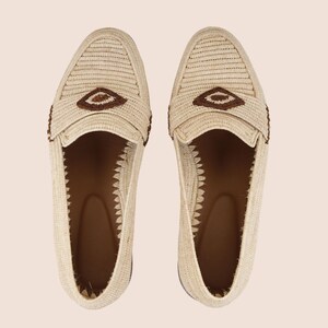 Puede incluir: Un par de mocasines beige tejidos con detalles marrones. Los zapatos presentan un dise&ntilde;o texturizado y un elemento decorativo marr&oacute;n en la parte superior. El interior es marr&oacute;n y los zapatos est&aacute;n sobre un fondo neutro.