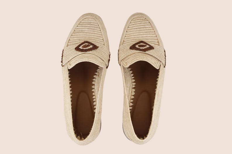 Puede incluir: Un par de mocasines beige tejidos con detalles marrones. Los zapatos presentan un dise&ntilde;o texturizado y un elemento decorativo marr&oacute;n en la parte superior. El interior es marr&oacute;n y los zapatos est&aacute;n sobre un fondo neutro.