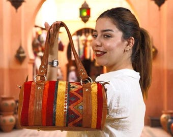 May include: A colorful handbag with a rectangular shape. The bag features a woven fabric design in red, orange, yellow, and blue, with brown leather accents and handles. The bag is held up by a person.