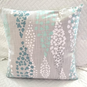 May include: Square decorative pillow with a gray background and a pattern of teal, white, and gray dots. The pillow has a textured appearance and is likely made of fabric. The design is abstract and modern.