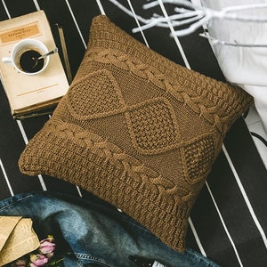 May include: A brown knitted throw pillow with a cable knit pattern. The pillow is square shaped and has a soft, cozy texture.