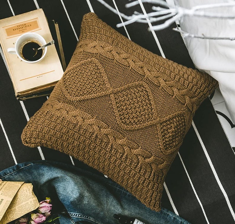 May include: A brown knitted throw pillow with a cable knit pattern. The pillow is square shaped and has a soft, cozy texture.