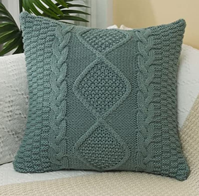 May include: A soft, green knitted throw pillow with a cable knit pattern. The pillow is square and has a diamond-shaped design in the center.