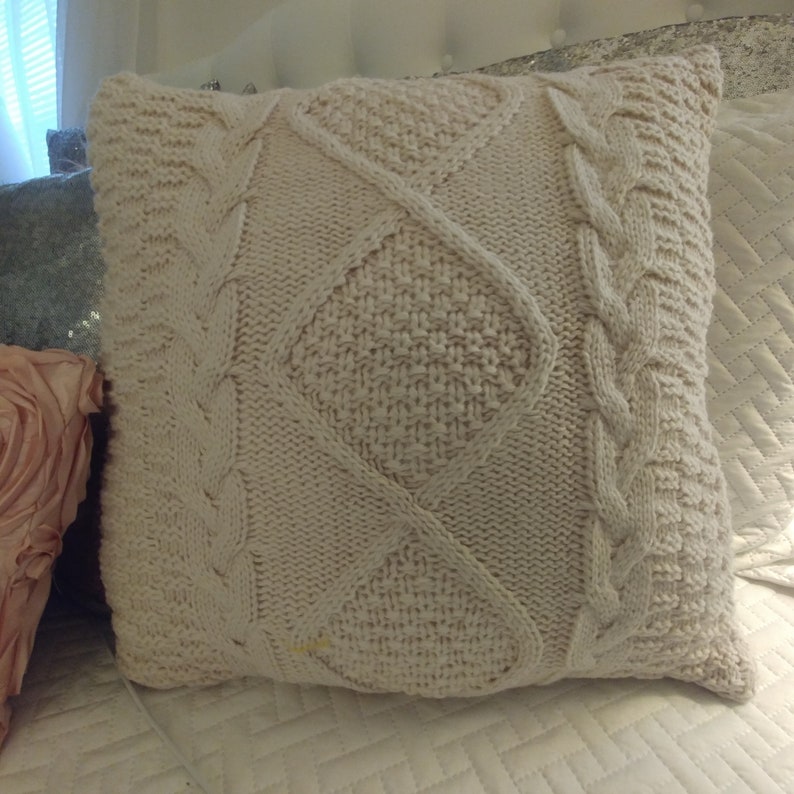 May include: A cream-colored knitted throw pillow with a cable knit pattern and diamond-shaped designs.