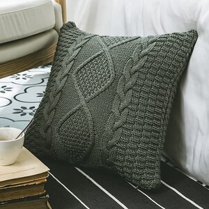 May include: A soft, gray knitted throw pillow with a cable knit pattern. The pillow is on a striped surface.