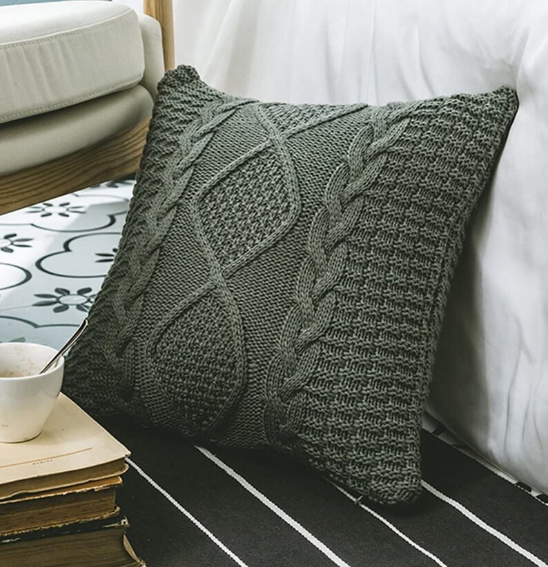 May include: A soft, gray knitted throw pillow with a cable knit pattern. The pillow is on a striped surface.
