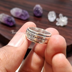 Peut inclure: Une bague rotative en argent dotée d'un anneau poli avec le texte gravé "I am enough" en écriture cursive. La bague possède une bande centrale rotative qui bouge indépendamment des bords extérieurs, présentant une finition métallique.
