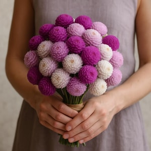 May include: A bouquet of vibrant, spherical flowers in shades of purple, lavender, and white. The blooms are tightly clustered, creating a full, textured appearance. The stems are bound with twine, held by a person wearing a gray dress.