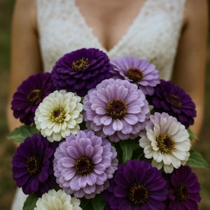 Op de afbeelding: Een boeket van zinnia's in tinten paars, lavendel en crème. De bloemen hebben gelaagde bloemblaadjes en donkere centra, met groen blad zichtbaar. Het boeket wordt tegen een vage achtergrond gehouden.