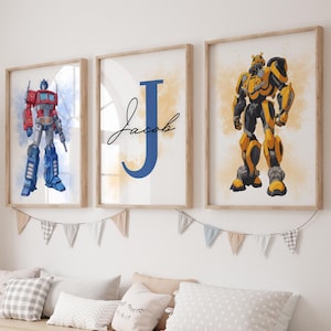 Può includere: Tre stampe da parete incorniciate che raffigurano un robot rosso e blu a sinistra, una grande lettera J blu con il nome Jacob in corsivo nero al centro e un robot giallo e nero a destra, il tutto all'interno di cornici in legno chiaro.