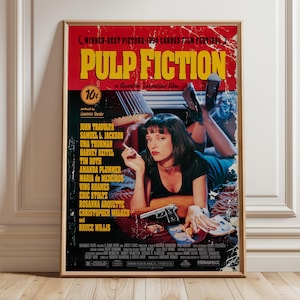 May include: A framed movie poster for "Pulp Fiction" with a red and yellow title. The poster features Uma Thurman, a gun, and the film's cast. The poster is displayed on a wooden floor against a white wall.