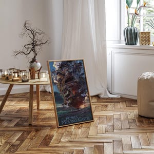 Op de afbeelding: Een ingelijste poster met de illustratie van Howl's Moving Castle. De afbeelding toont het kasteel in grijstinten, bruin en rood, tegen een blauwe lucht en een groen landschap. De tekst "Howl's Moving Castle" staat onderaan.