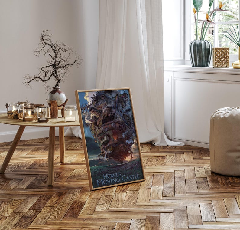 Op de afbeelding: Een ingelijste poster met de illustratie van Howl's Moving Castle. De afbeelding toont het kasteel in grijstinten, bruin en rood, tegen een blauwe lucht en een groen landschap. De tekst "Howl's Moving Castle" staat onderaan.