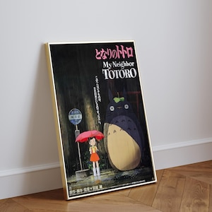May include: A framed poster featuring the animated film "My Neighbor Totoro." The artwork depicts a young girl holding a red umbrella, standing near a bus stop with Totoro in a rainy forest setting. Japanese text is visible.
