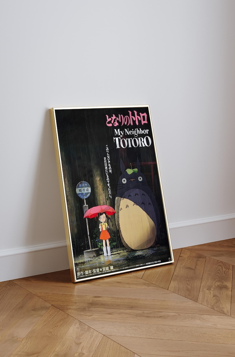May include: A framed poster featuring the animated film "My Neighbor Totoro." The artwork depicts a young girl holding a red umbrella, standing near a bus stop with Totoro in a rainy forest setting. Japanese text is visible.