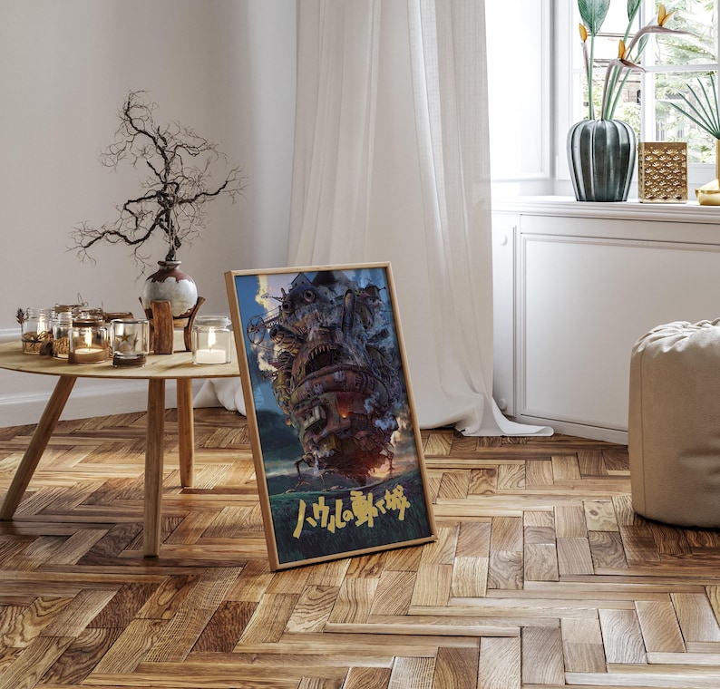 May include: A framed poster featuring a detailed illustration of a moving castle with smoke billowing from its top. The artwork is set against a blue sky and green landscape. Japanese text is visible at the bottom of the poster. The poster is displayed on a wooden floor.