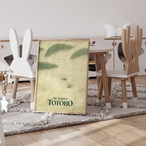 May include: A framed print of "My Neighbor Totoro" is propped against a wall. The artwork features a light green and yellow background with two small figures and green foliage. The print is displayed in a child's room with a white bunny-shaped chair.