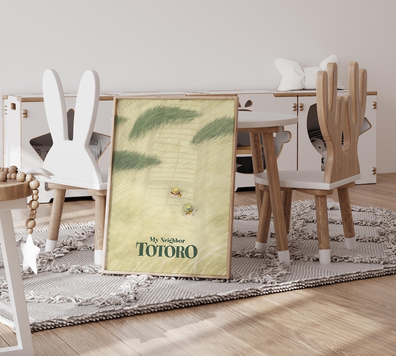 May include: A framed print of "My Neighbor Totoro" is propped against a wall. The artwork features a light green and yellow background with two small figures and green foliage. The print is displayed in a child's room with a white bunny-shaped chair.