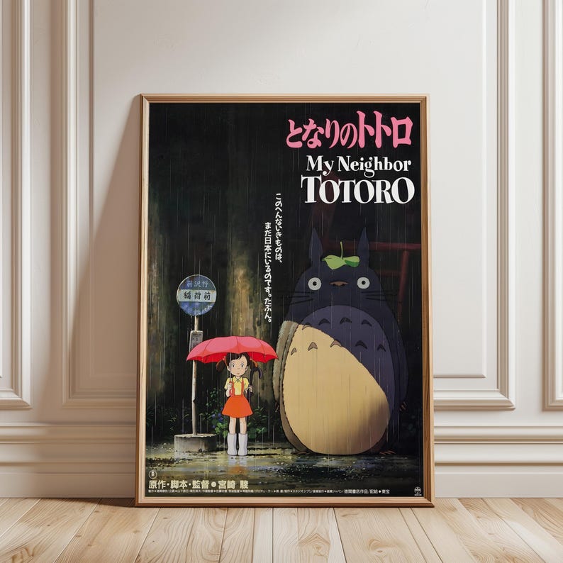 May include: A framed poster featuring the animated film "My Neighbor Totoro." The artwork depicts a young girl with a red umbrella standing at a bus stop with Totoro, a large forest spirit. The poster has Japanese text.