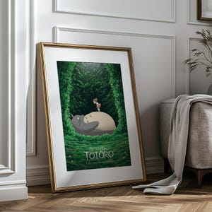 May include: Framed art print featuring a scene from "My Neighbor Totoro." The artwork depicts Totoro, a large gray forest spirit, with a young girl standing on his belly. The background is a lush green forest, with the title "TOTORO" at the bottom.