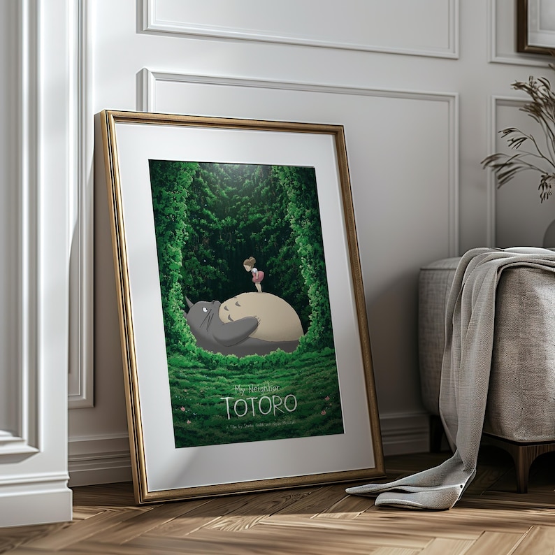 May include: Framed art print featuring a scene from "My Neighbor Totoro." The artwork depicts Totoro, a large gray forest spirit, with a young girl standing on his belly. The background is a lush green forest, with the title "TOTORO" at the bottom.