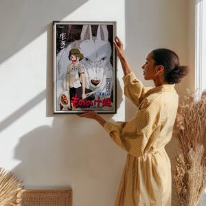 May include: Framed poster featuring anime artwork. The image showcases a girl with a wolf, with Japanese text. The poster has a black frame and is being held up against a white wall. The artwork is colorful, with a focus on the characters and their expressions.